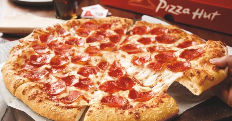 large pepperoni pizza hut pizza on table with person taking a slice off