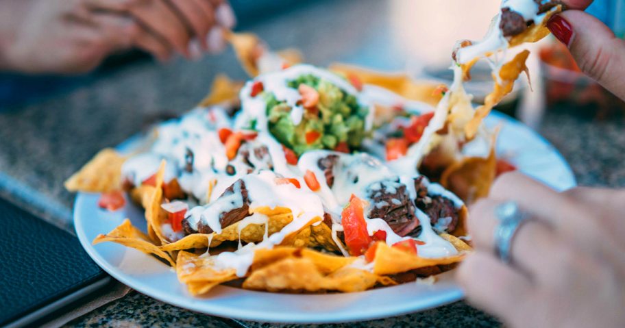 plate full of nachos on table with peoples hands picking chips