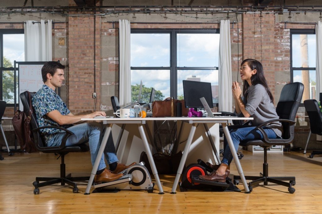 man at woman sitting at opposite sides of desk both using elliptical 