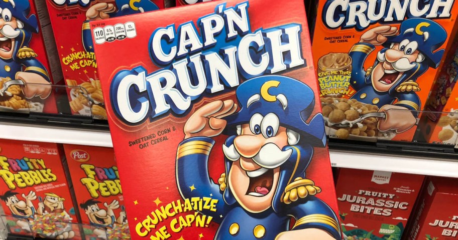 hand holding a box of Cap'n Crunch Cereal with more boxes on a store shelf behind it