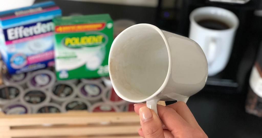 hand holding a white stained coffee mug with denture tablet boxes in the background