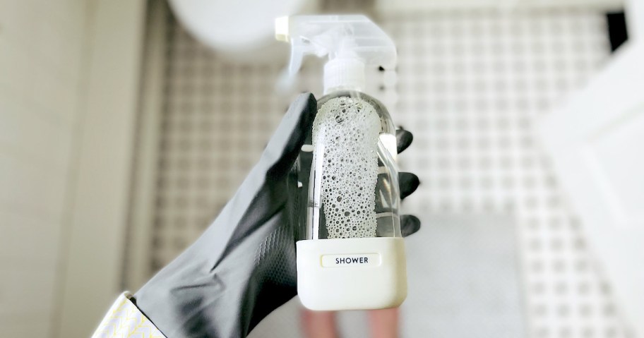 hand holding a clear spray bottle with white silicone sleeve labeled shower on bottom