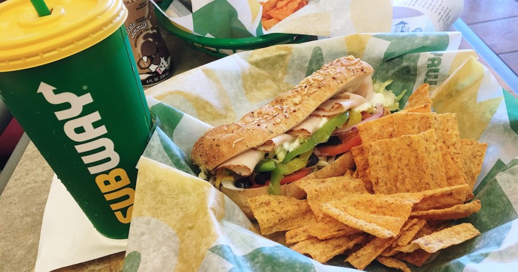 green subway cup next to plate with chips and half of a sub sandwich