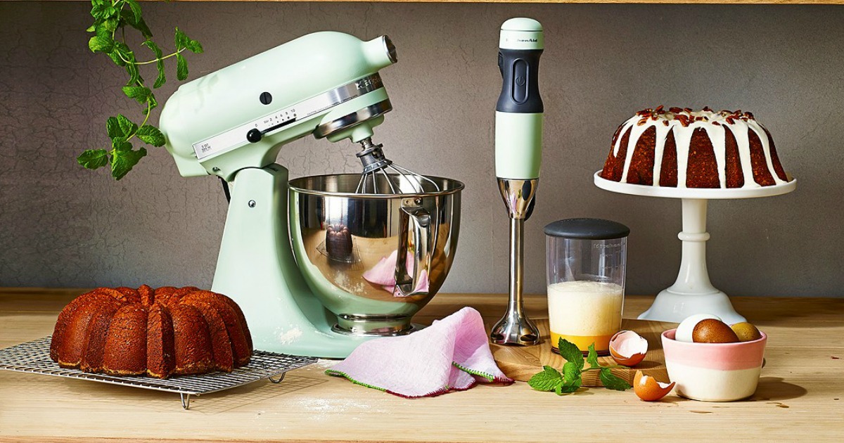 light green Kitchenaid mixer on wooden counter next to immersion blender and several baked goods