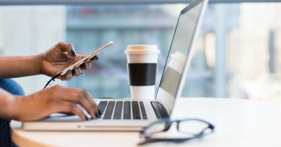 person typing on laptop while looking at information on a cellphone