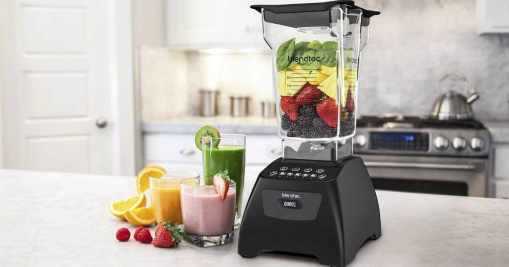 Black Blendtec-brand blender on a counter top filled with fruits and vegetables, with prepared smoothies nearby