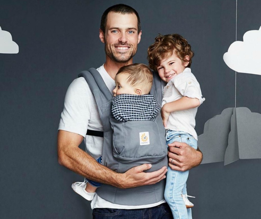 Father wearing baby in a carrier while holding toddler with a gray background 