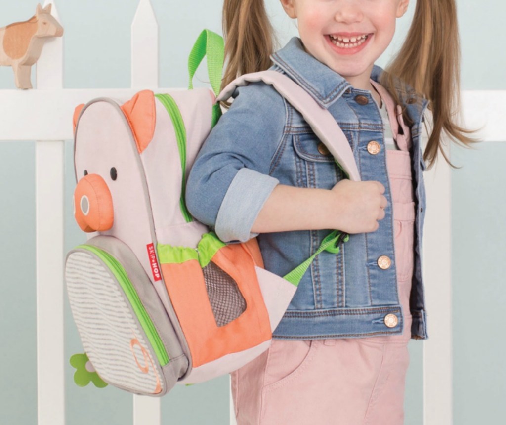 Girl wearing SkipHop brand Pig themed backpack near white picket fence