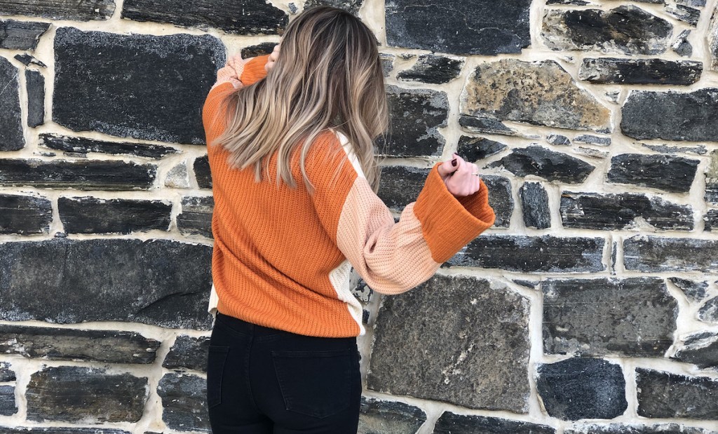 woman wearing orange colorblock sweater facing stone wall 