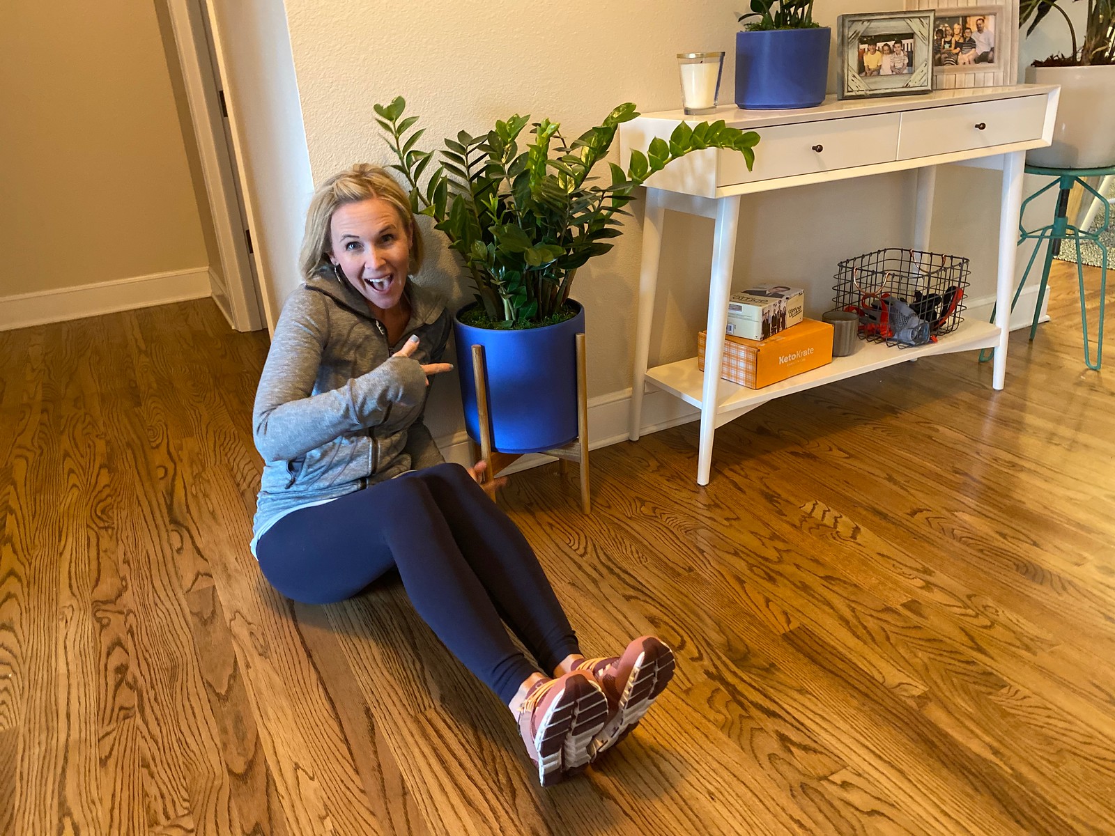 woman on the ground in home pointing at large potted plant 