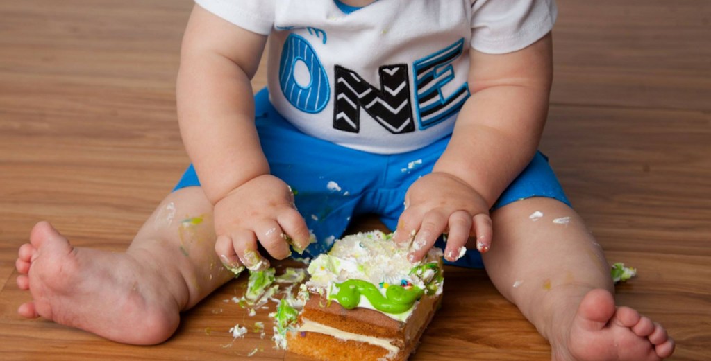 baby with one shirt sitting on wood floor touching piece of birthday cake