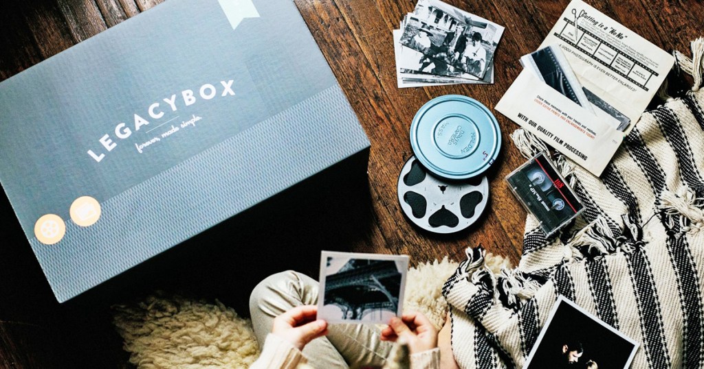 person holding old photo in hand with more photos, film reel, and legacybox in background on wood floor