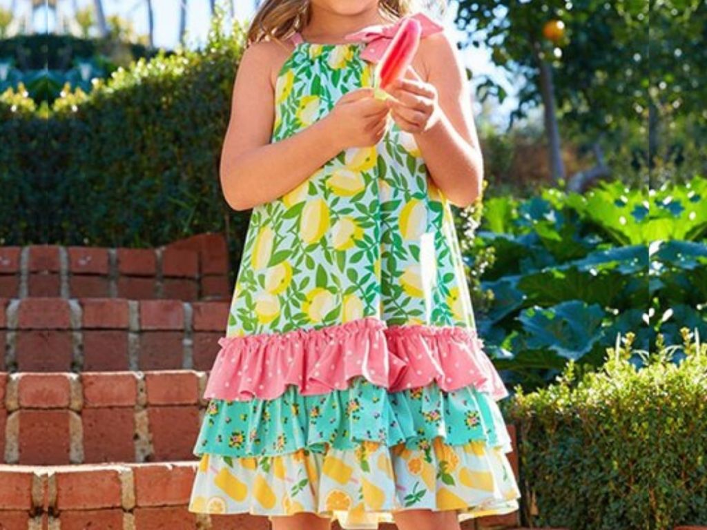 little girl wearing frilled summer dress, eating a popsicle on front of brick stairs