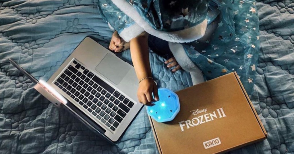 child wrapped in blue blanket sitting on bed in front of laptop and coding kit