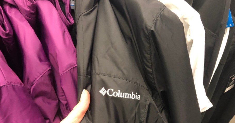 person's hand on a black Columbia jacket, more jackets in other colors around it on a clothes rack