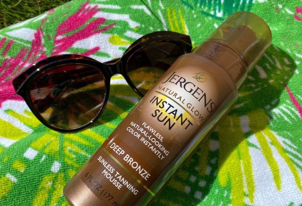 bottle of jergens self tanner and sunglasses laying on green beach towel