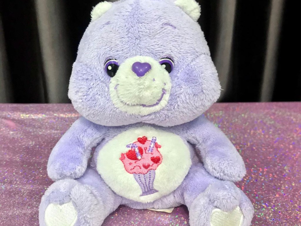 purple plush bear with hearts and milkshake design on stomach on purple tablecloth