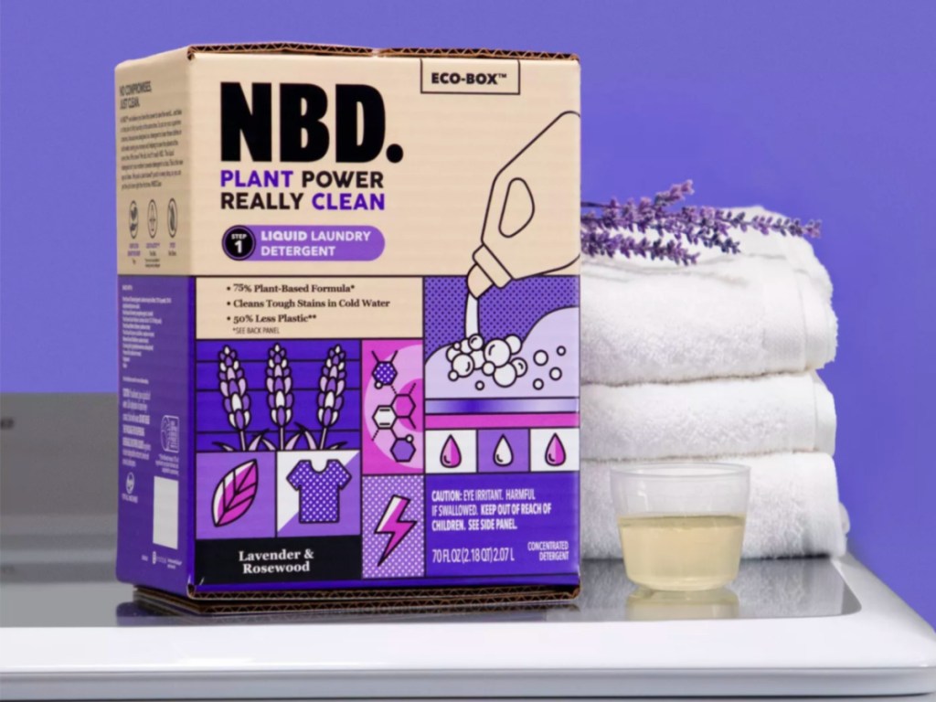 box of NBD Lavender & Rosewood Liquid Laundry Detergent with cup fill of laundry detergent and 3 folded white towels