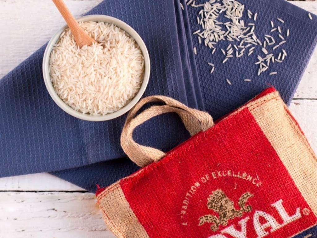 Large bag of rice with handles near small bowl of rice on white wooden surface