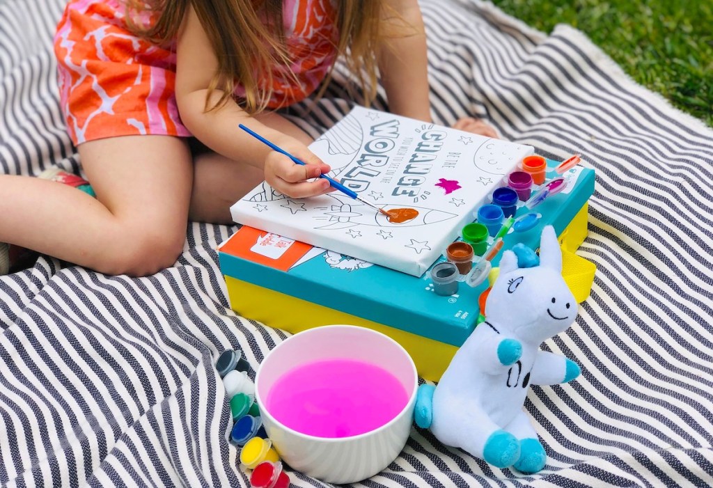 girl painting on blank canvas outside on blanket in grass