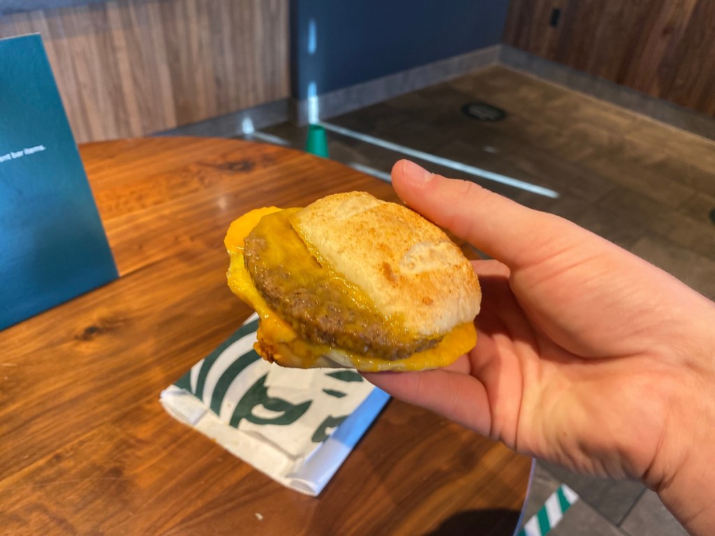 hand holding sausage egg and cheese sandwich with Starbucks bag on table nearby