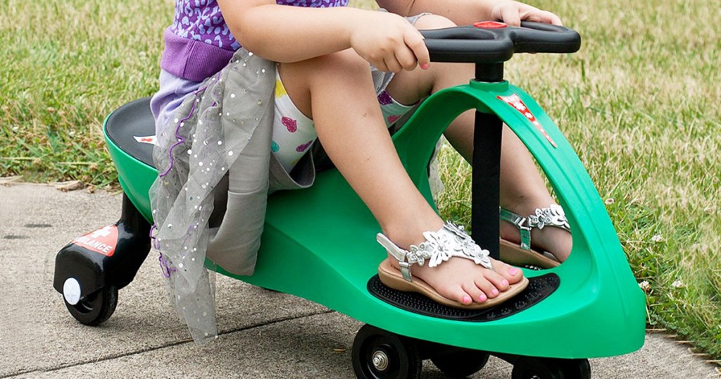 girl wearing grey skirt and sandals sitting on a green wiggle car on sidewalk