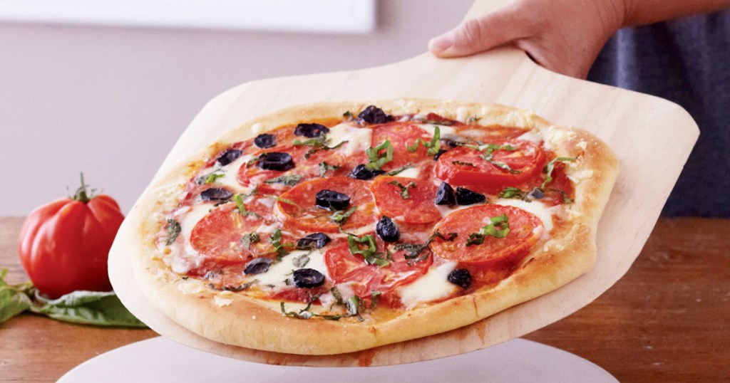 person holding a wood pizza paddle with a pizza on top