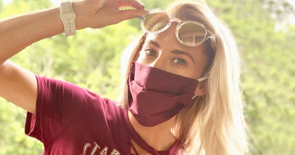 woman in red shirt wearing a matching red face mask standing outside holding up a pair of sunglasses on her face