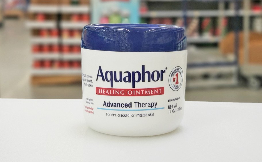 large white and blue 14oz jar of Aquaphor Healing Ointment on white table, store shelves in the background blurred out