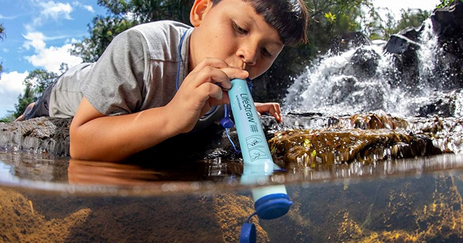 boy laying down to stick blue lifestraw into stream to drink