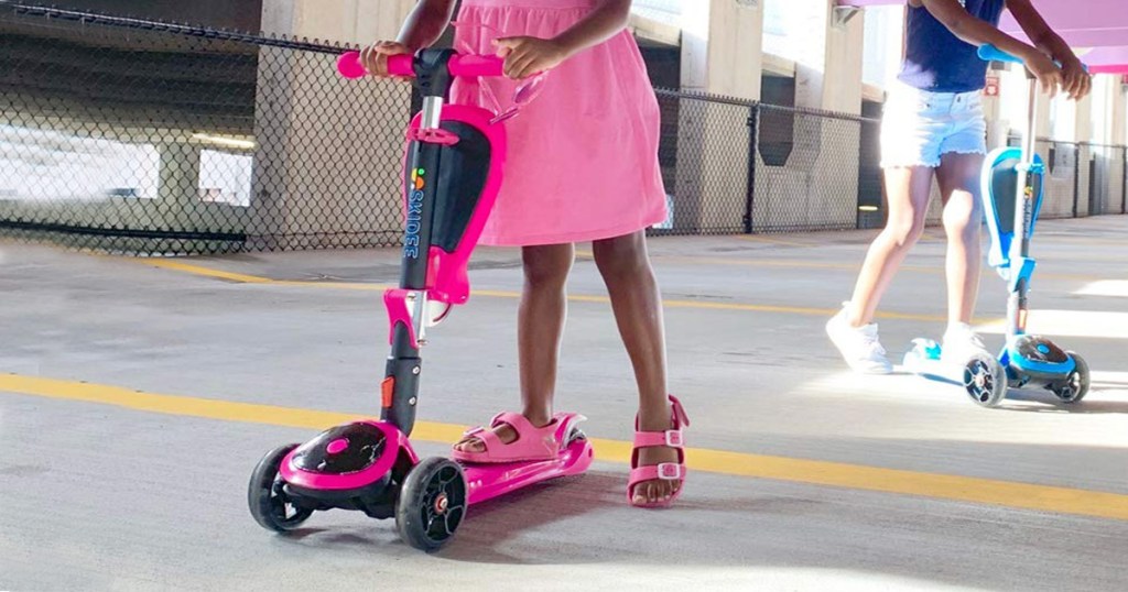 two girls riding on 3 wheeled scooters in parking garage