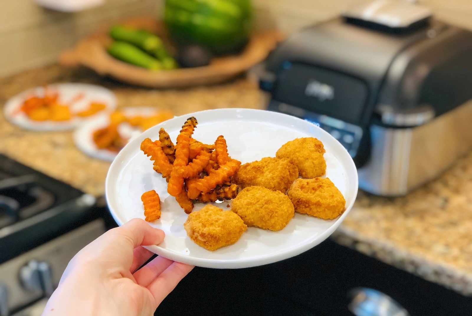 hand holding white plate with fries and nuggets in front of ninja foodi grill