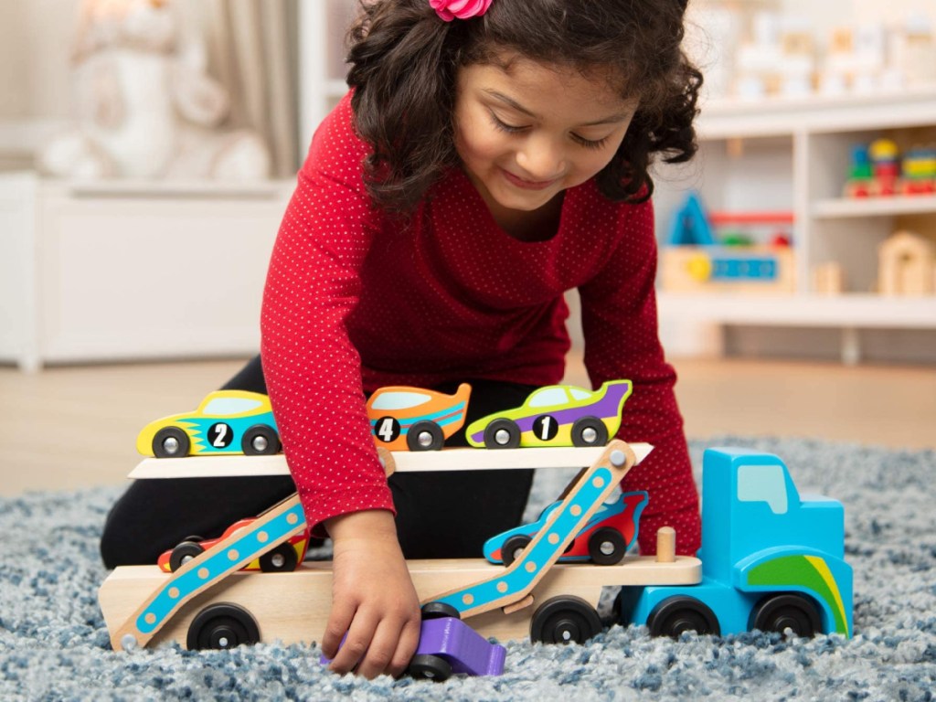 little girl kneeling on the floor playing with a wooden car carrier toy