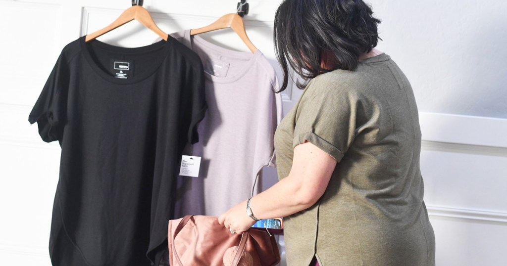 woman with black hair wearing olive green shirt standing next to other tunic tops hanging from wooden hangers