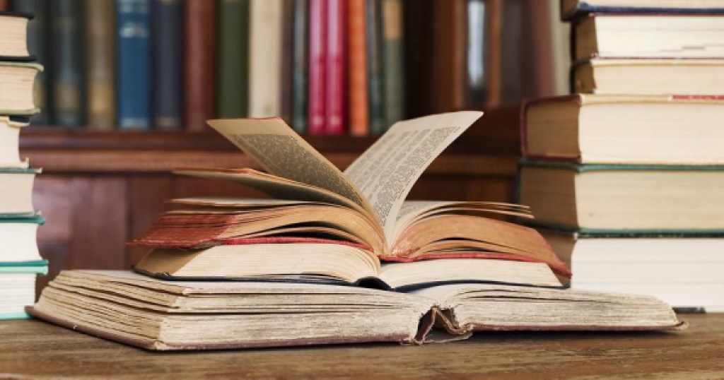 multiple books stacked up on shelfs and table with a small pile in the middle with one open