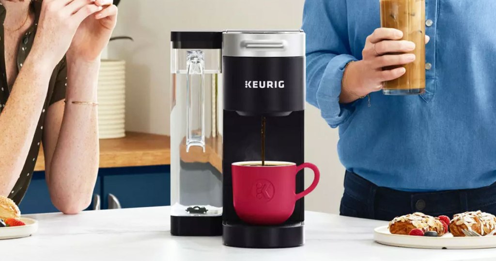 black keurig coffee maker brewing into red cup with people behind it in kitchen