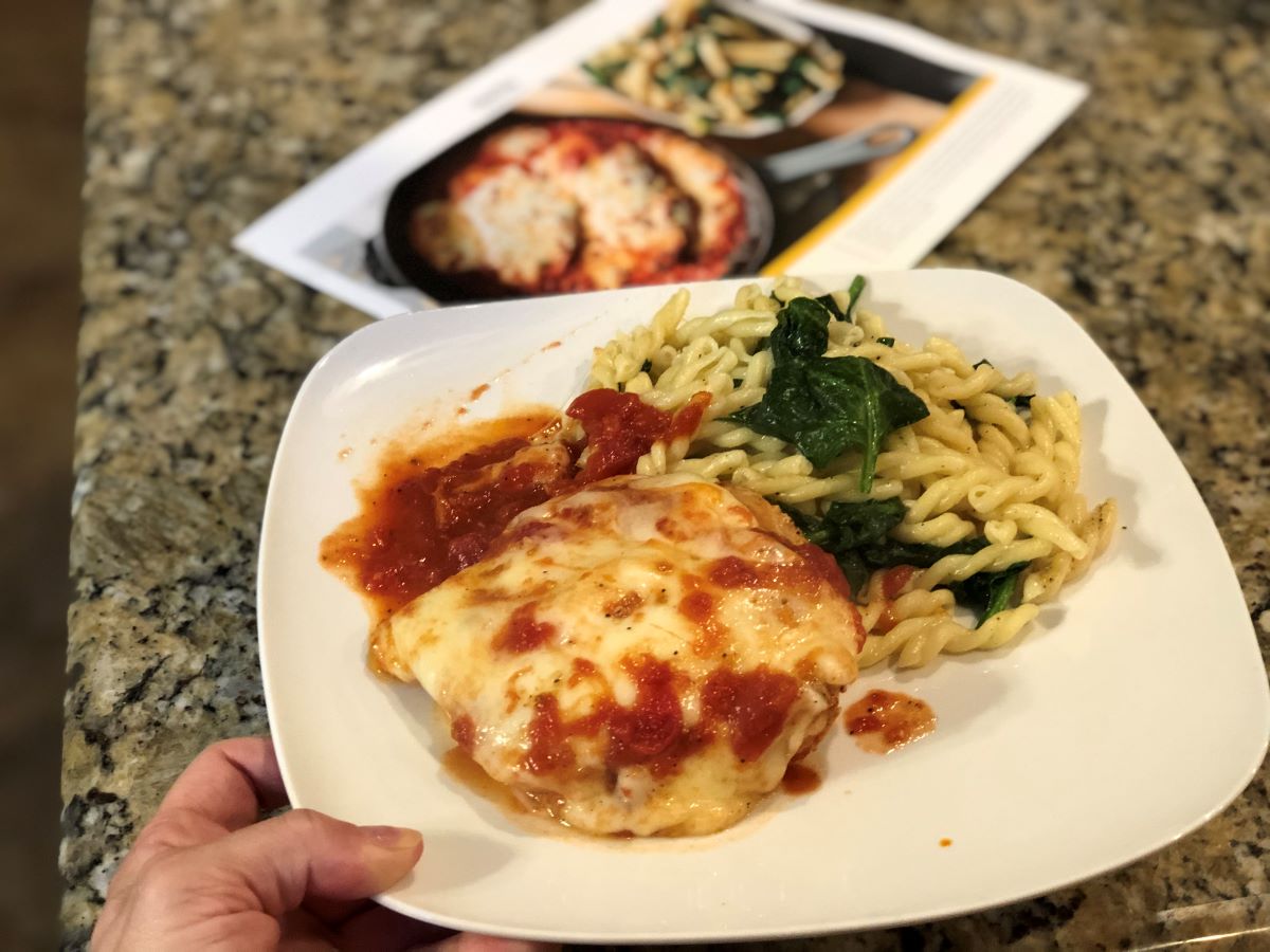 hand holding a plate with cheesy chicken and pasta on it