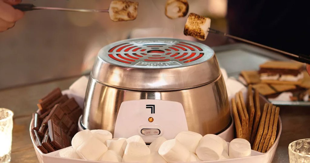 three people roasting marshmallows over a stainless steel smores maker with ingredients in tray around it