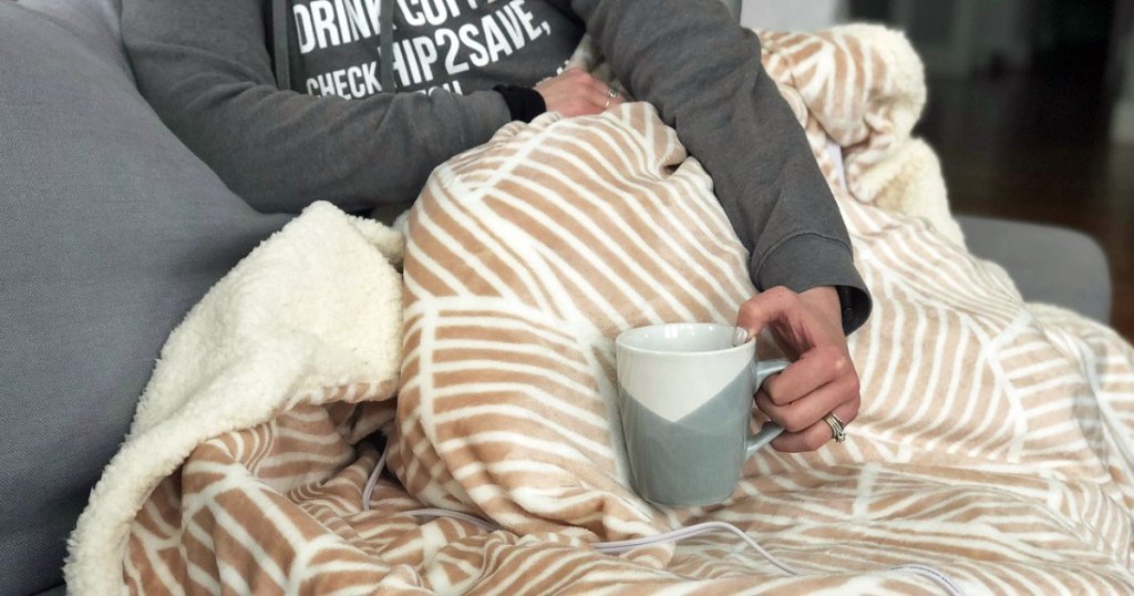 woman in grey sweatshirt under an electric blanket holding a coffee mug