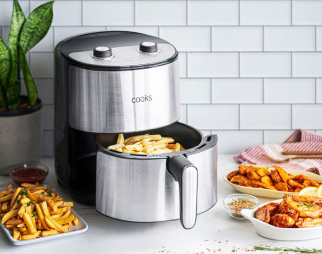 round stainless steel air fryer on kitchne counter with fries in basket and plates of fried foods around it