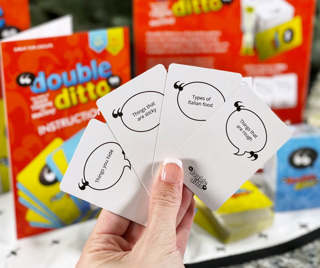 woman holding up four playing cue cards from the double ditto family game with the red box and more playing cards in the background