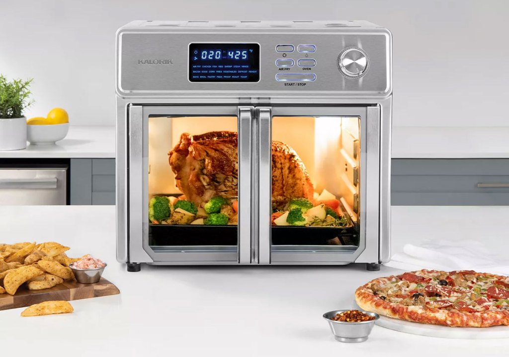 stainless steel air fryer oven on kitchen counter with roast chicken inside and pizza and fries on plates in front of it