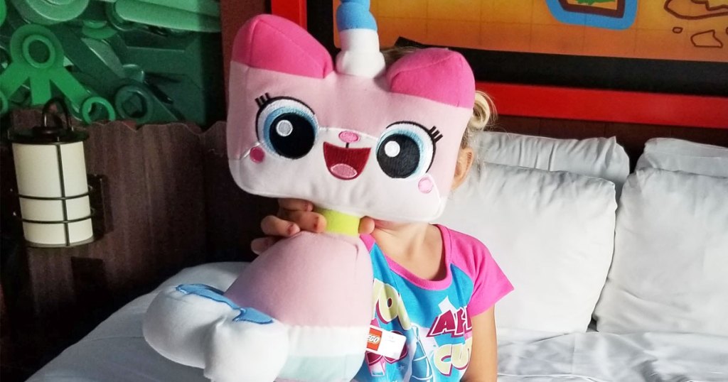 girl sitting on a bed holding up a pink lego movie unikitty plush