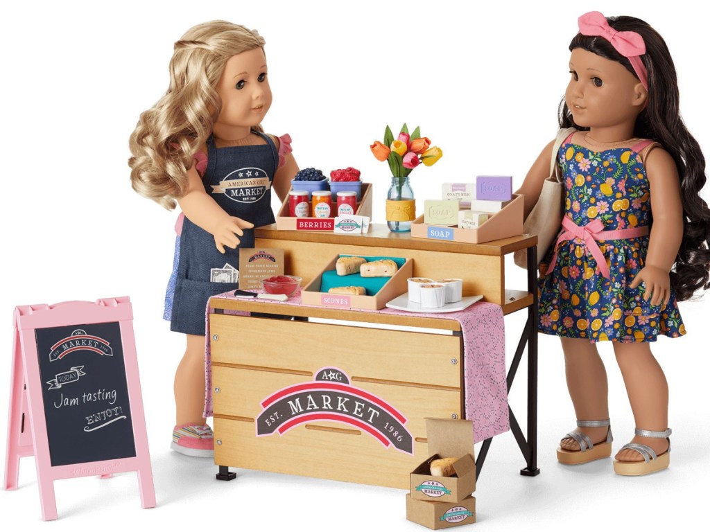 2 american girl dolls standing next to a market playset