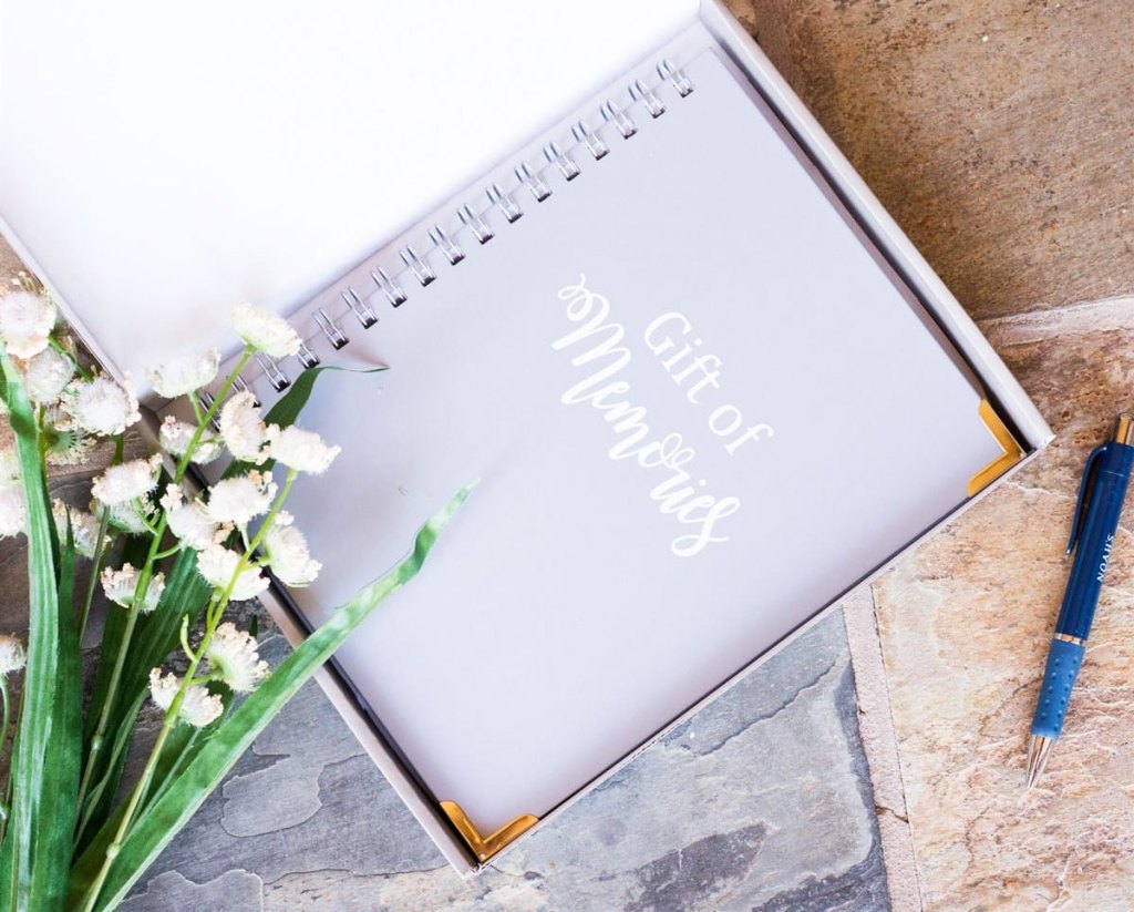 spiral bound notebook that says gift of memories with flowers and pen near it