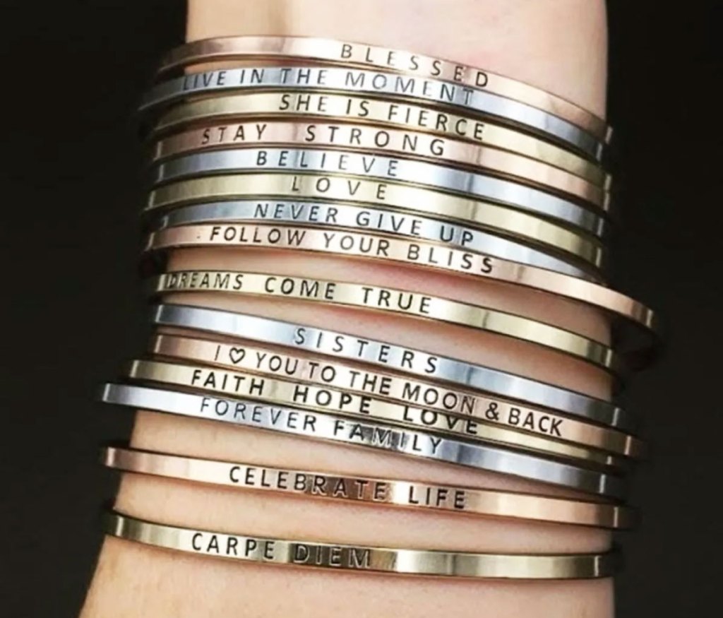 woman's wrist with a ton of thin metal bracelets with inspirational signs on them in gold, silver, and rose gold colors