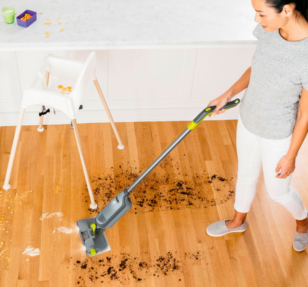 woman in grey shirt and white pants using a grey shark vacmop to clean up dirt and crumbs off hardwood floor