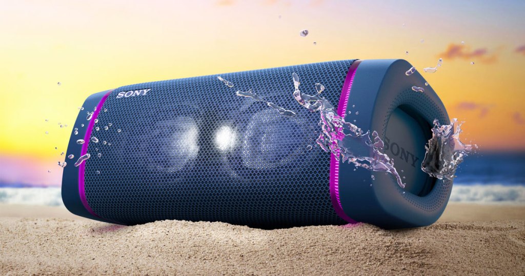 navy blue sony portable speaker with purple led lights on either side in sand with water splashes around it