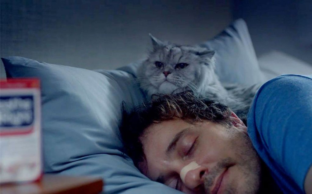 man in bed sleeping with breathe right nasal strip on his nose and cat laying at his head