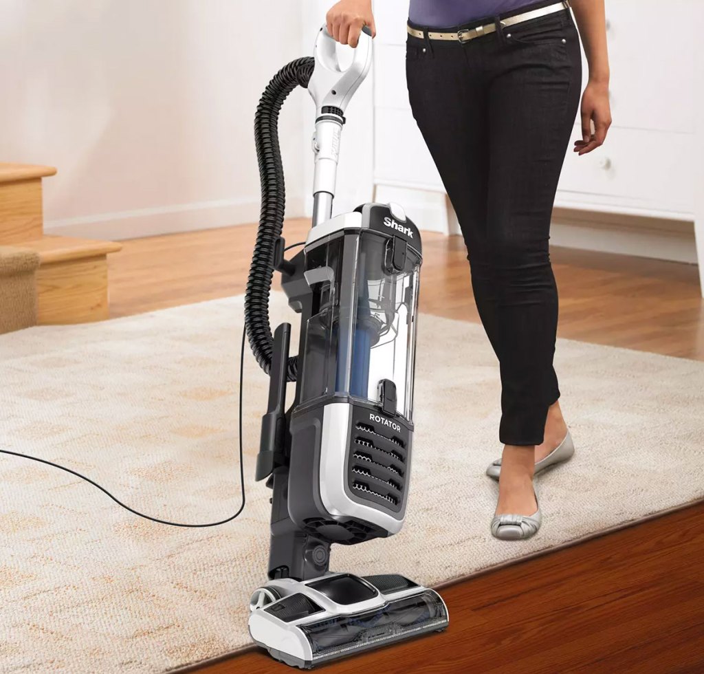 woman in black pants pushing a grey and white shark upright vacuum on area rug